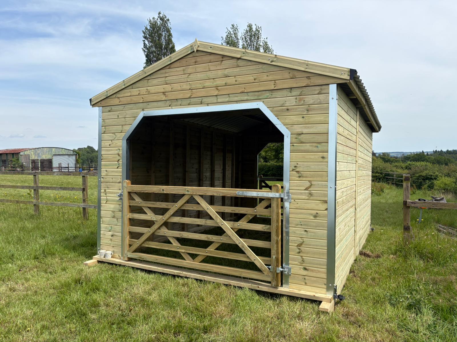 Premium 12 x 18 walk through horse shelter featuring timber cladding, steel-reinforced frame, and gated access at both ends in a grass field setting.